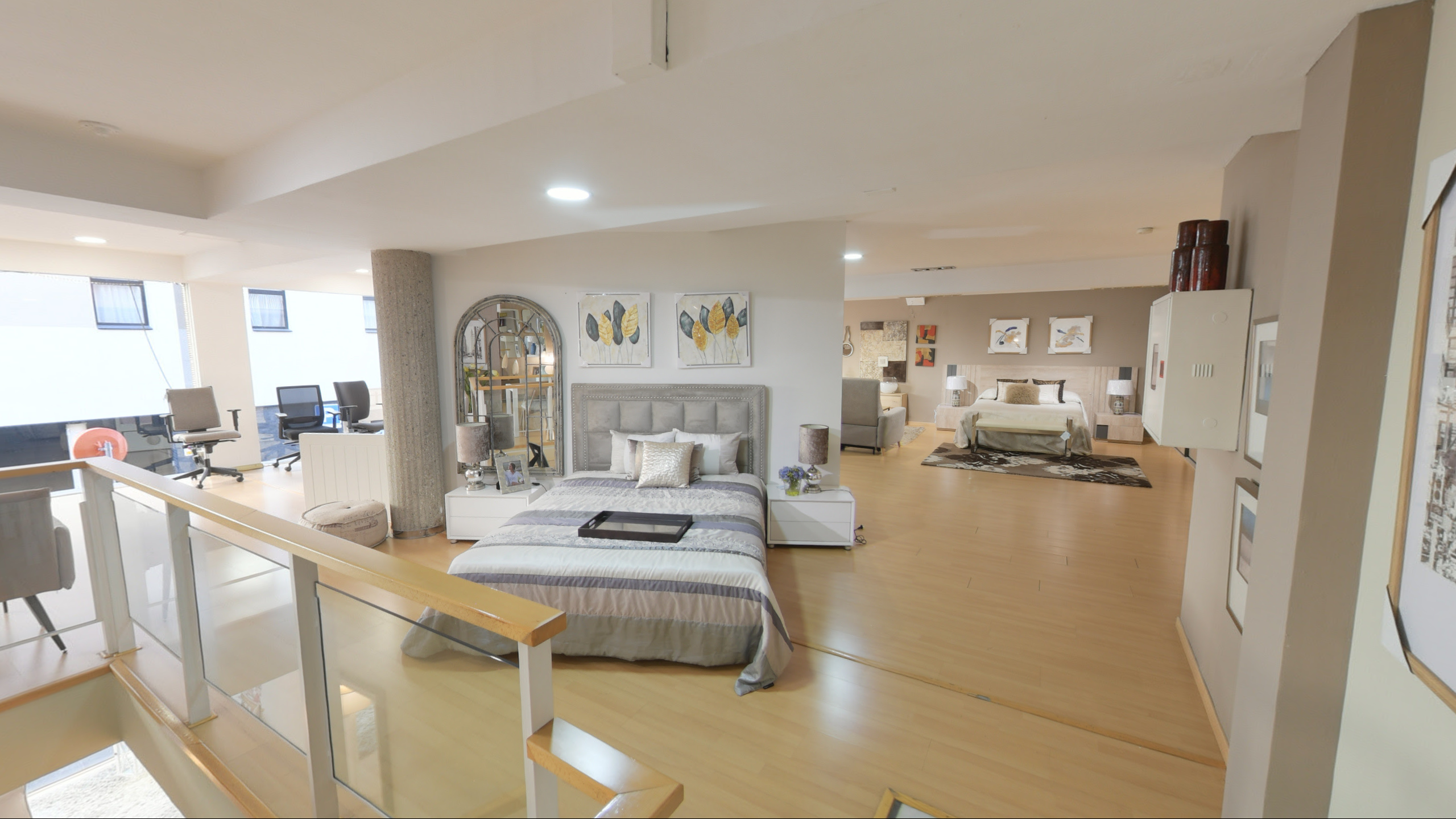 Tienda de Muebles en Barcelona, mobiliario a medida y diseño - Temas Mobiliario Exposición de dormitorio con cama de matrimonio, cabecero tapizado en gris con capitoné, mesitas de noche blancas y cuadros decorativos en la pared.