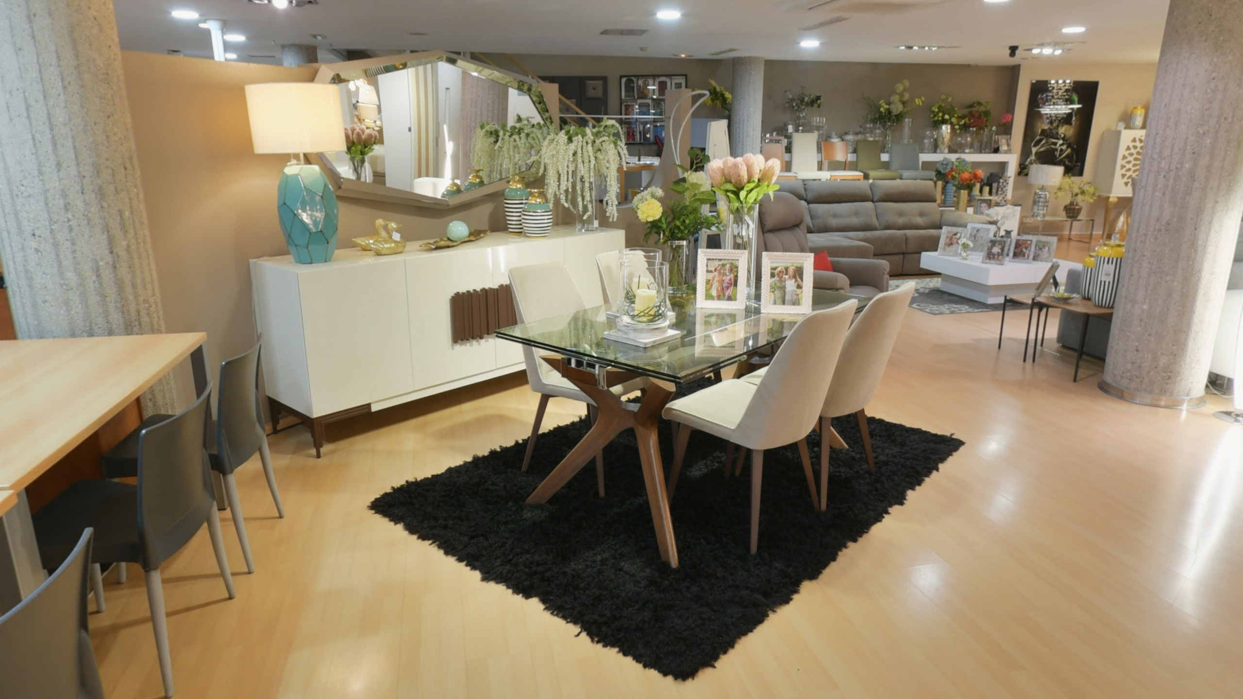 Tienda de Muebles en Barcelona, mobiliario a medida y diseño - Temas Mobiliario Conjunto de comedor moderno con mesa de sobre de cristal y patas de madera, sillas tapizadas en beige, aparador blanco y un gran espejo geométrico al fondo