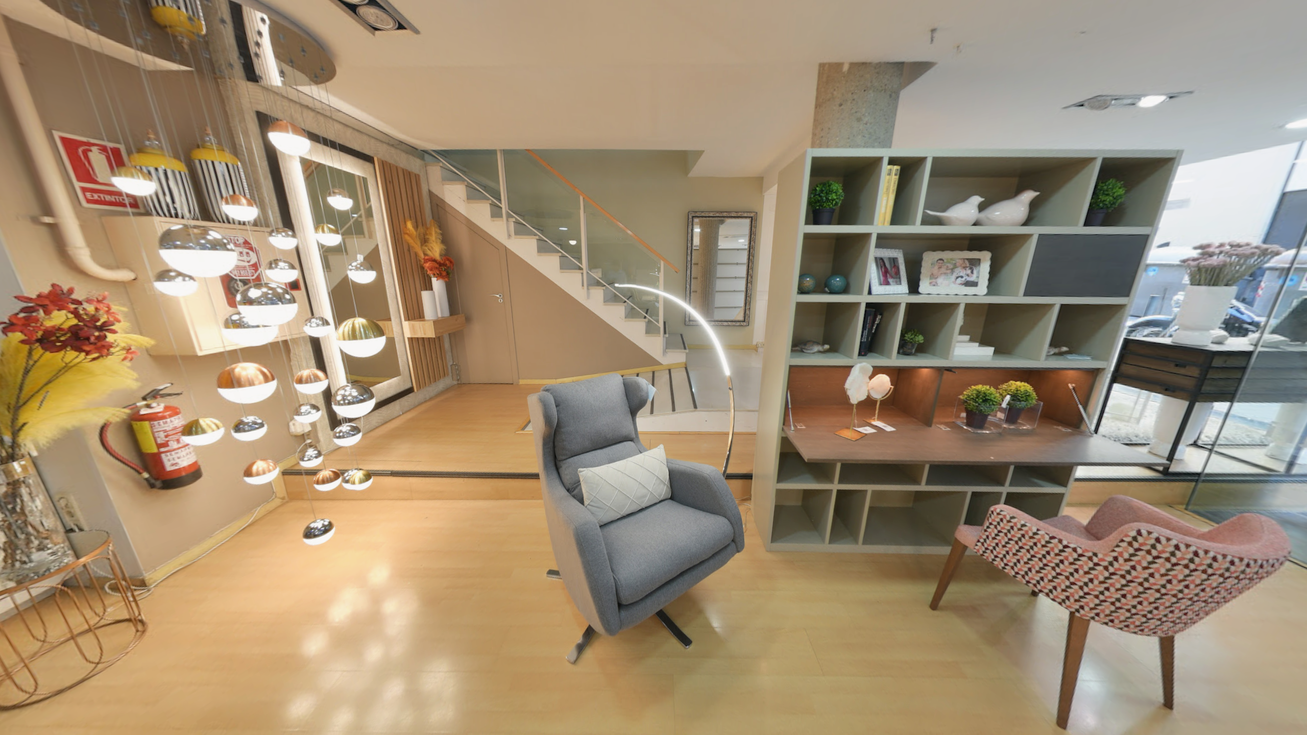 Tienda de Muebles en Barcelona, mobiliario a medida y diseño - Temas Mobiliario Espacio de diseño que incluye una estantería-librería en tono gris oliva con escritorio integrado, una butaca giratoria gris con cojín blanco y una silla de acento con estampado geométrico. Al fondo se observa una escalera con barandilla de cristal y una moderna lámpara de cascada con múltiples globos metálicos.